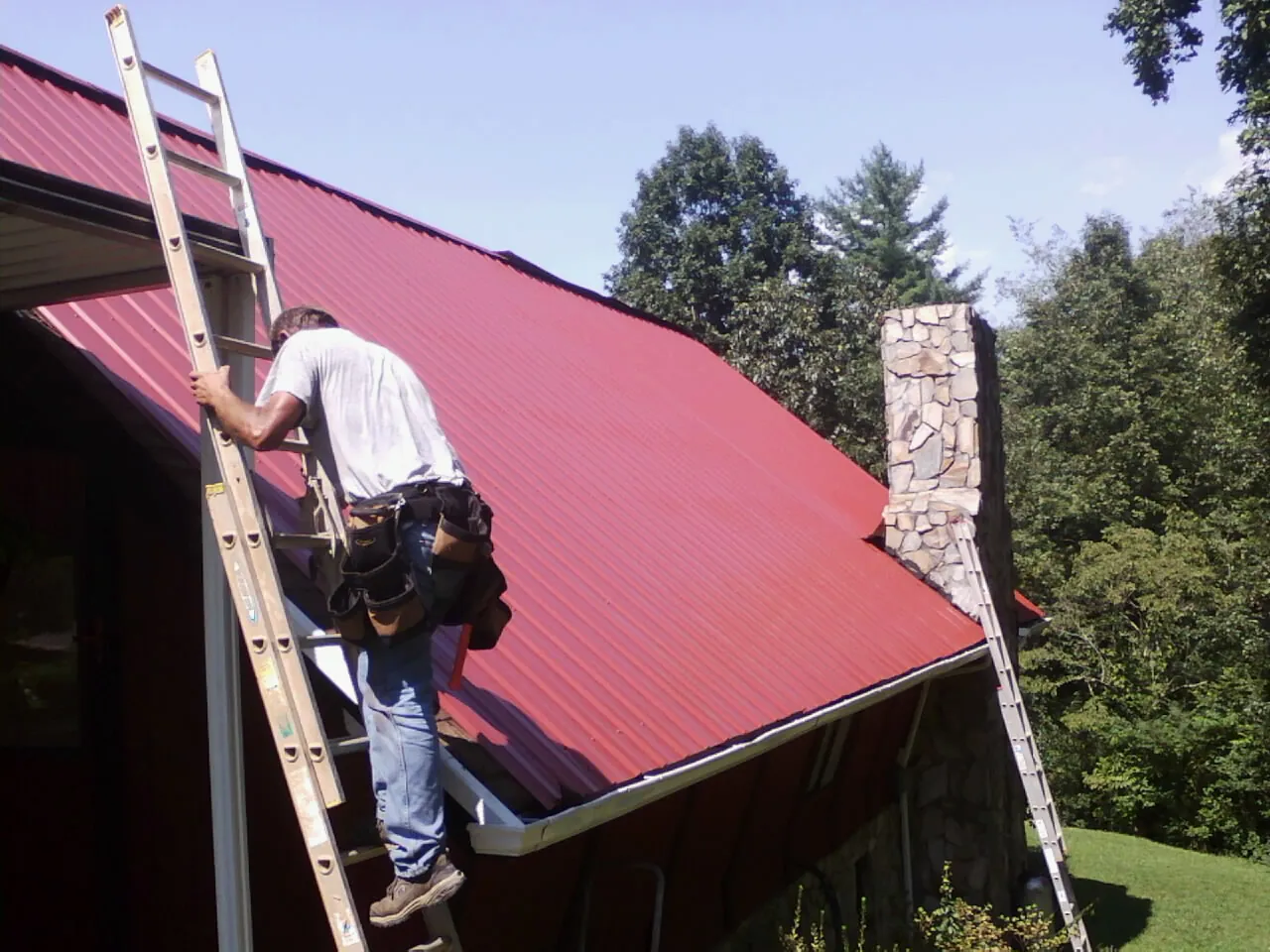 Roofer on a ladder beside a red metal roof and stone chimney — Soffit & Fascia Repair by Roofing Brothers in Bear River City